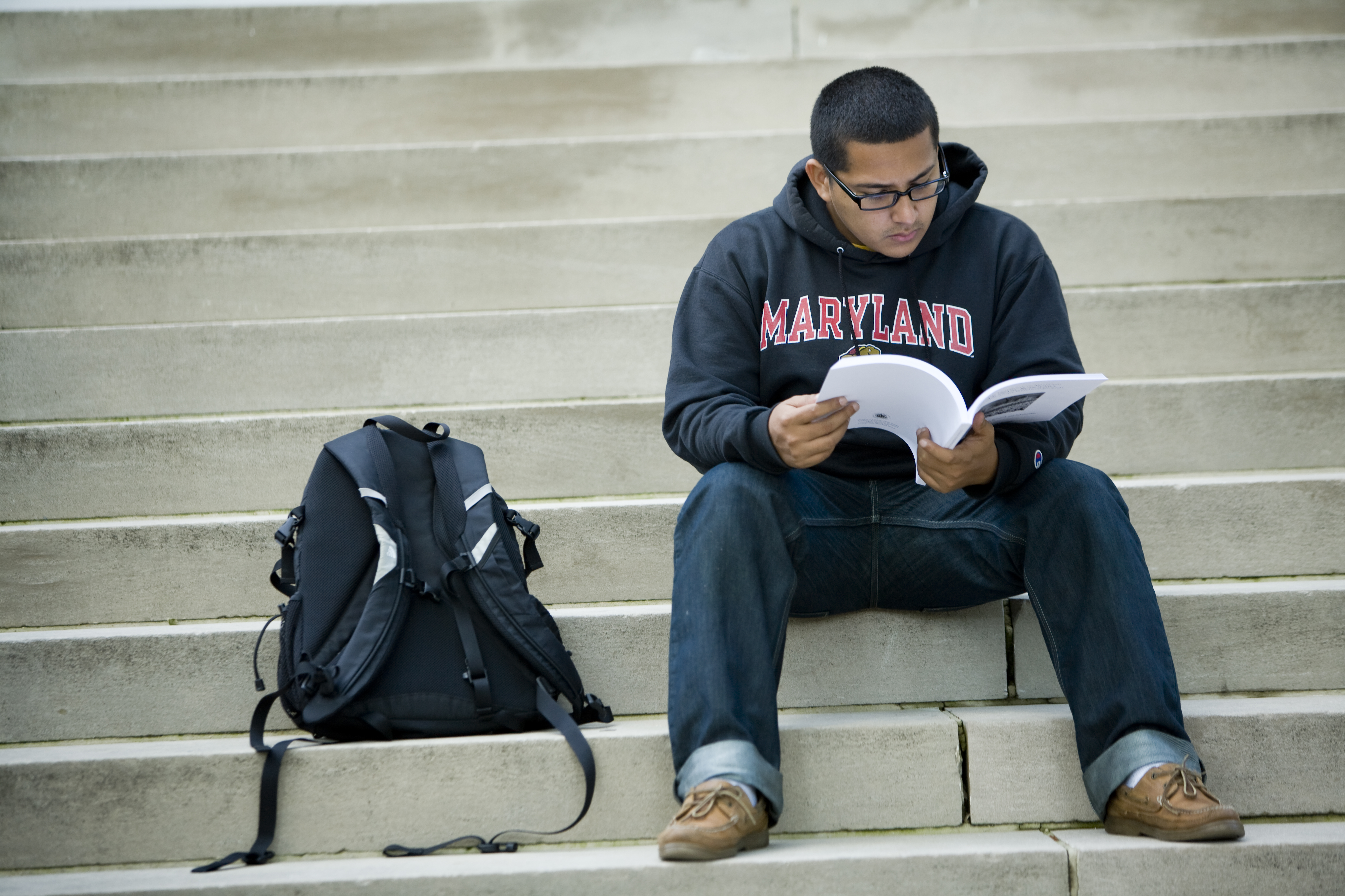 student in m shirt sitting on step with open book